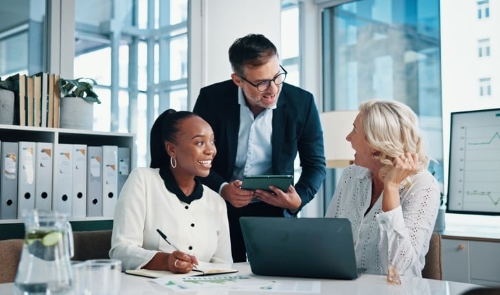 A team of three professionals meets in a bright office, smiling and collaborating over sales and finance data on laptops and printed reports.