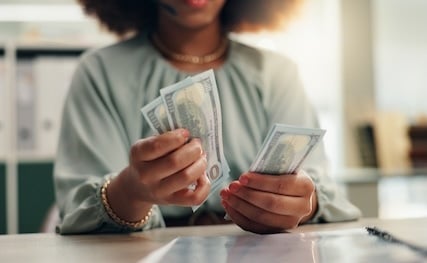 Close-up of a businesswoman counting U.S. hundred-dollar bills at her desk, symbolizing sales commissions, variable pay, or incentive compensation.