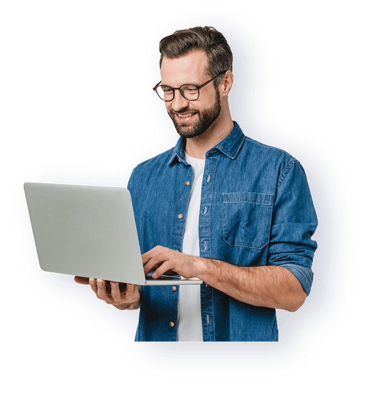 A man in a denim shirt and glasses is smiling while using a laptop