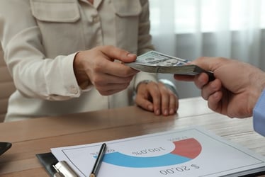 Close-up of a businessperson handing over cash across a desk with a financial chart in the foreground, symbolizing variable compensation and performance-based pay.