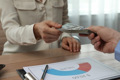 Close-up of a businessperson handing over cash across a desk with a financial chart in the foreground, symbolizing variable compensation and performance-based pay.