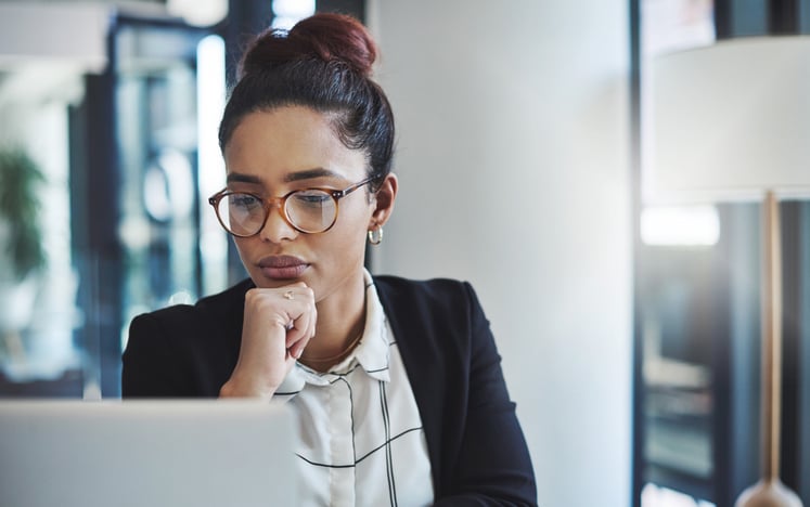 Woman in glasses and a blazer studies her laptop screen, chin resting on her hand, in a bright office